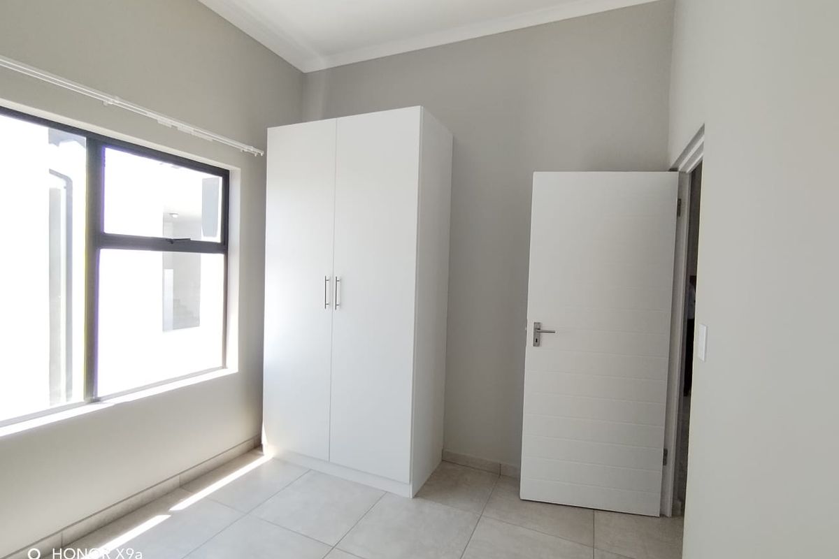 A bright, empty bedroom featuring neutral tiled flooring, a built-in white wardrobe, and modern aluminium window frames.