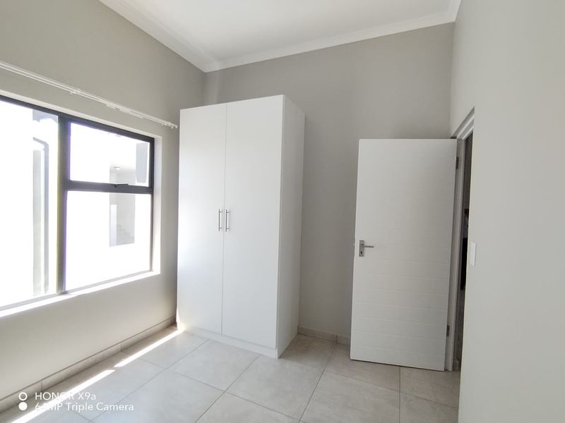 A bright, empty bedroom featuring neutral tiled flooring, a built-in white wardrobe, and modern aluminium window frames.