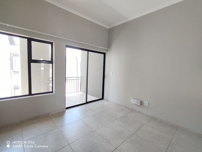 A bright, empty living area featuring neutral tiled flooring, large aluminium-framed windows, and a sliding glass door leading to an outdoor space.