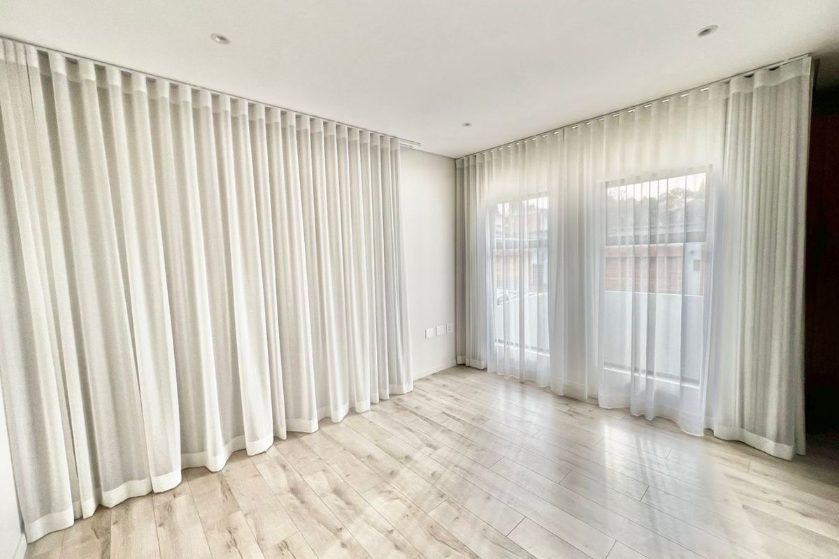 A bright, empty living room featuring light-toned laminate flooring, recessed ceiling lighting, and floor-to-ceiling sheer curtains.