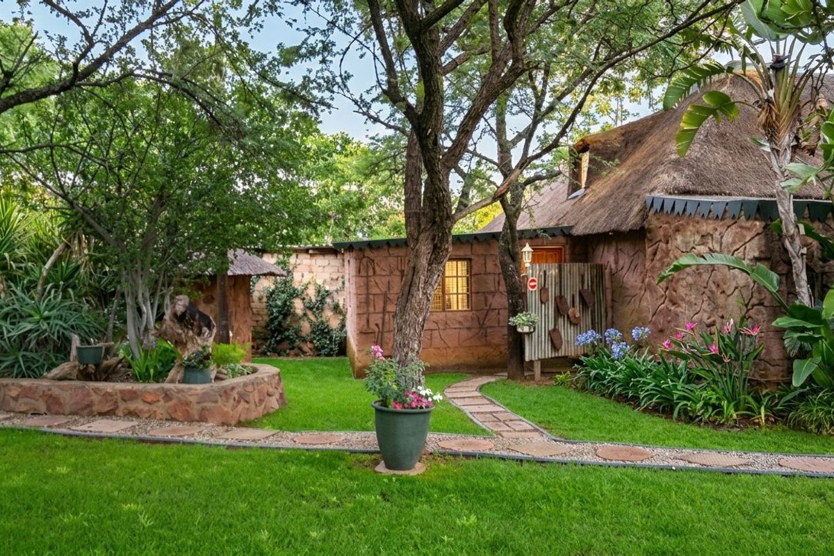 A charming rear garden area featuring a rustic cottage with a traditional thatch roof, stone cladding, and a well-maintained lawn with stone pathways.