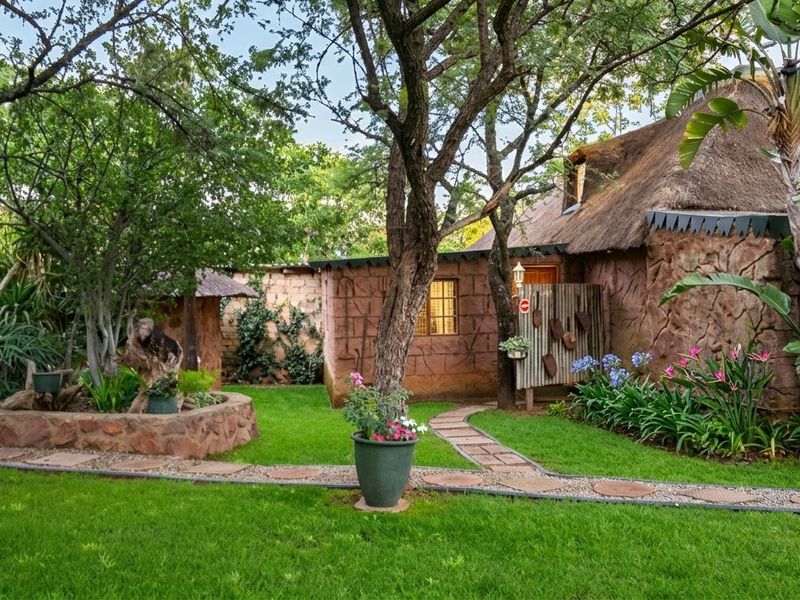 A charming rear garden area featuring a rustic cottage with a traditional thatch roof, stone cladding, and a well-maintained lawn with stone pathways.