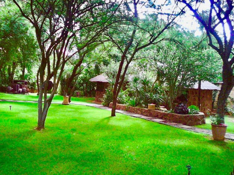 A lush, well-maintained garden area featuring mature trees, a stone retaining wall, and a paved pathway winding through the green lawn.