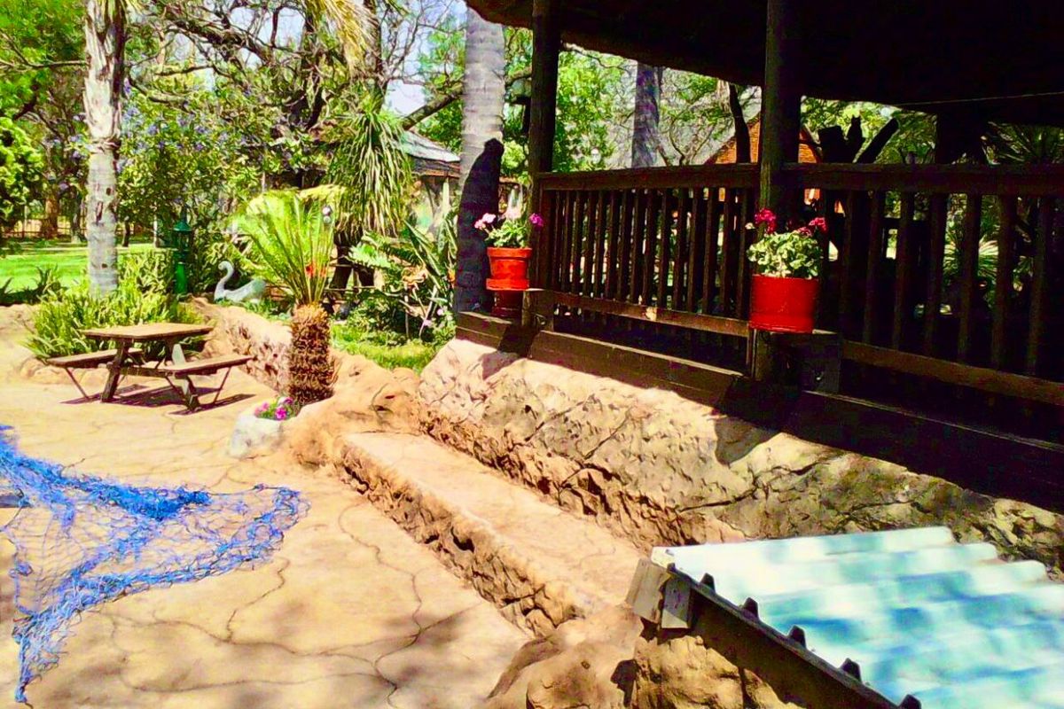 A rustic outdoor patio area featuring a thatched roof, timber balustrade, and stone-clad retaining wall, set within a lush garden environment.