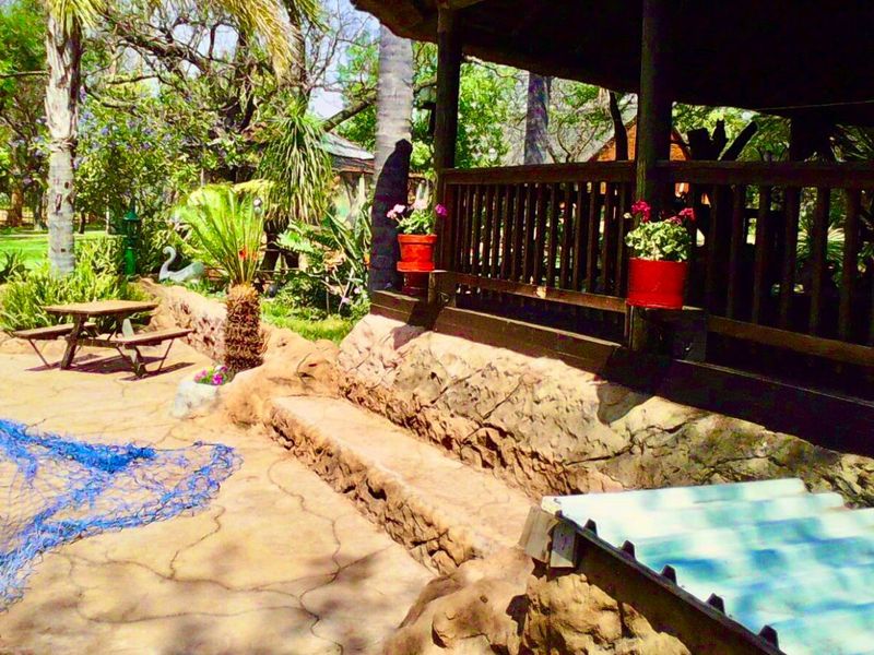 A rustic outdoor patio area featuring a thatched roof, timber balustrade, and stone-clad retaining wall, set within a lush garden environment.