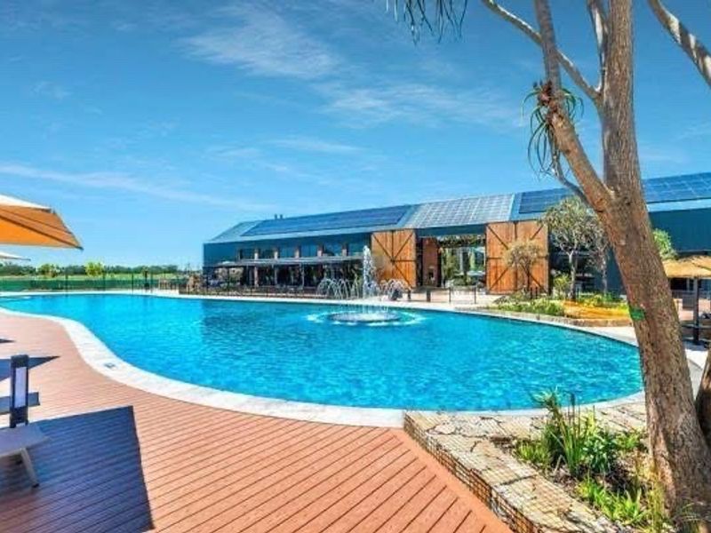 A well-maintained outdoor pool area featuring a large swimming pool with a central fountain, surrounded by timber decking and modern building architecture.