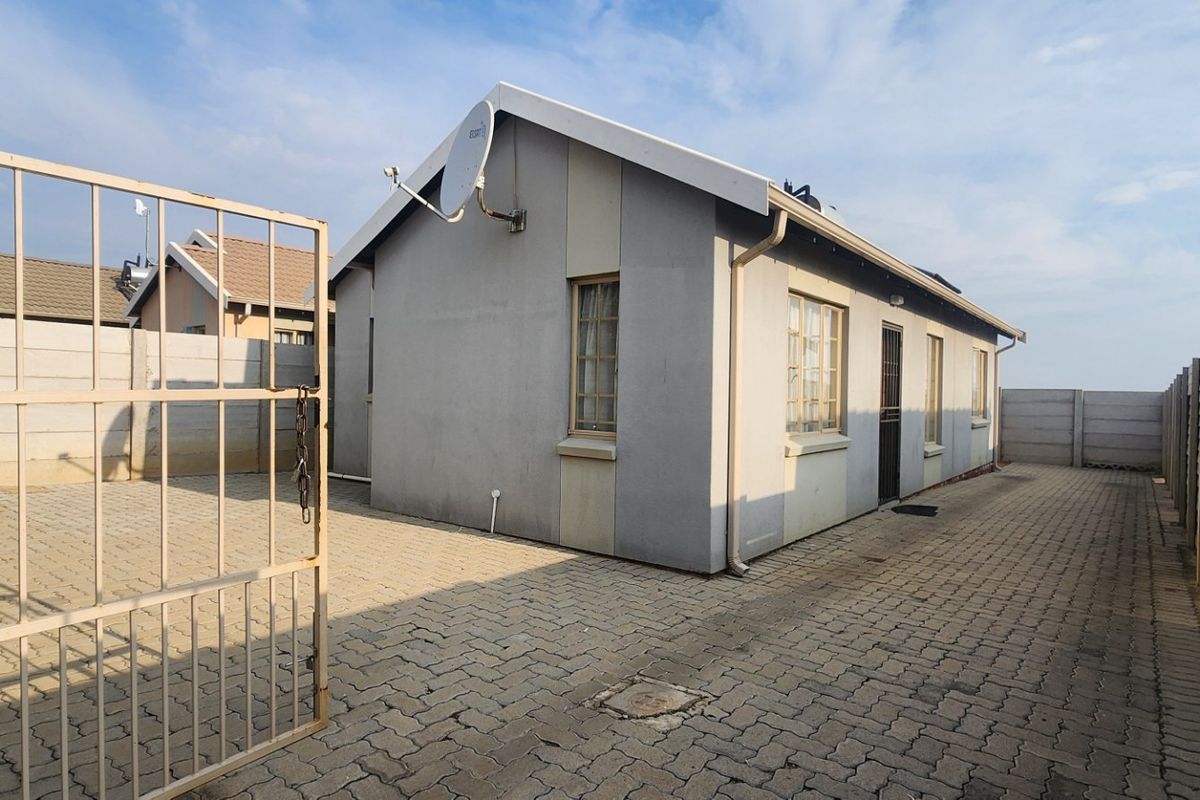 Rear exterior view of the property featuring a paved courtyard, secure metal gate, and precast concrete boundary walls.