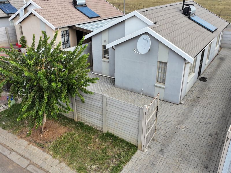 Aerial view of the front exterior featuring a paved driveway, precast boundary wall, metal gate, and a tiled roof with a solar water heater.