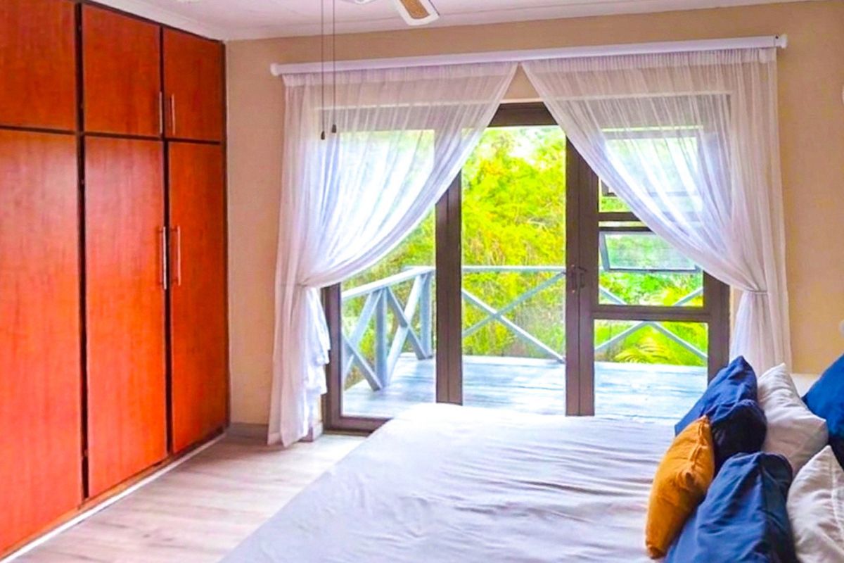 A bright bedroom featuring built-in wooden cupboards, laminate flooring, and sliding glass doors that open onto a private balcony with lush greenery views.