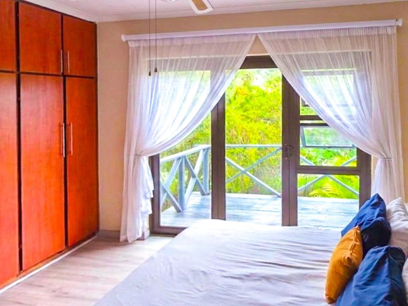 A bright bedroom featuring built-in wooden cupboards, laminate flooring, and sliding glass doors that open onto a private balcony with lush greenery views.