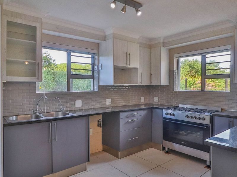 A functional kitchen featuring grey and light wood cabinetry, granite countertops, a gas stove, and a tiled backsplash with ample natural light.