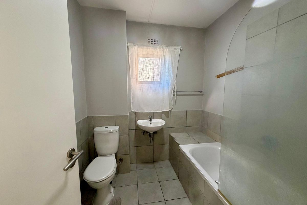 A functional bathroom featuring a built-in bathtub with a glass shower screen, a wall-mounted basin, and a toilet, finished with neutral floor and wall tiles.
