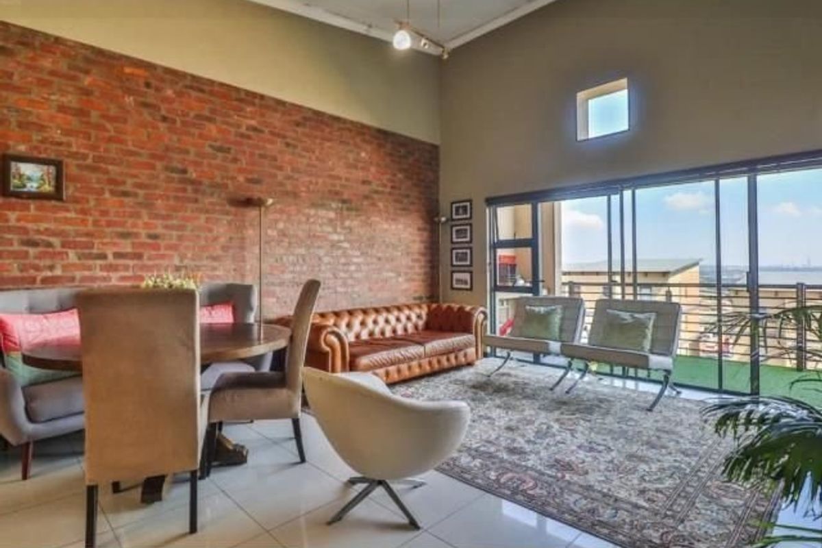 Spacious open-plan living area featuring a striking exposed brick wall, high vaulted ceilings, and large sliding doors leading to an outdoor balcony.
