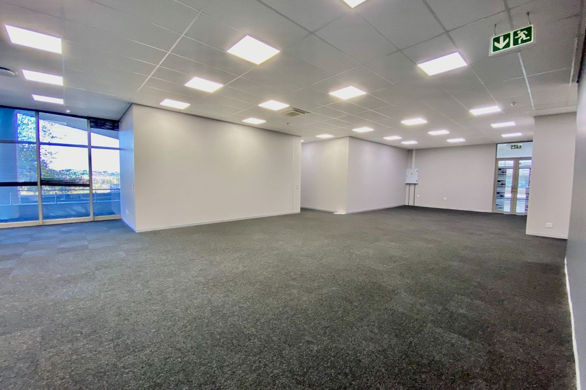 A spacious, vacant commercial office area featuring neutral finishes, carpeted flooring, recessed lighting, and large windows providing natural light.