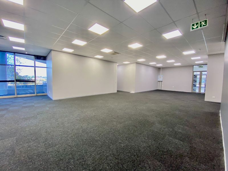 A spacious, vacant commercial office area featuring neutral finishes, carpeted flooring, recessed lighting, and large windows providing natural light.