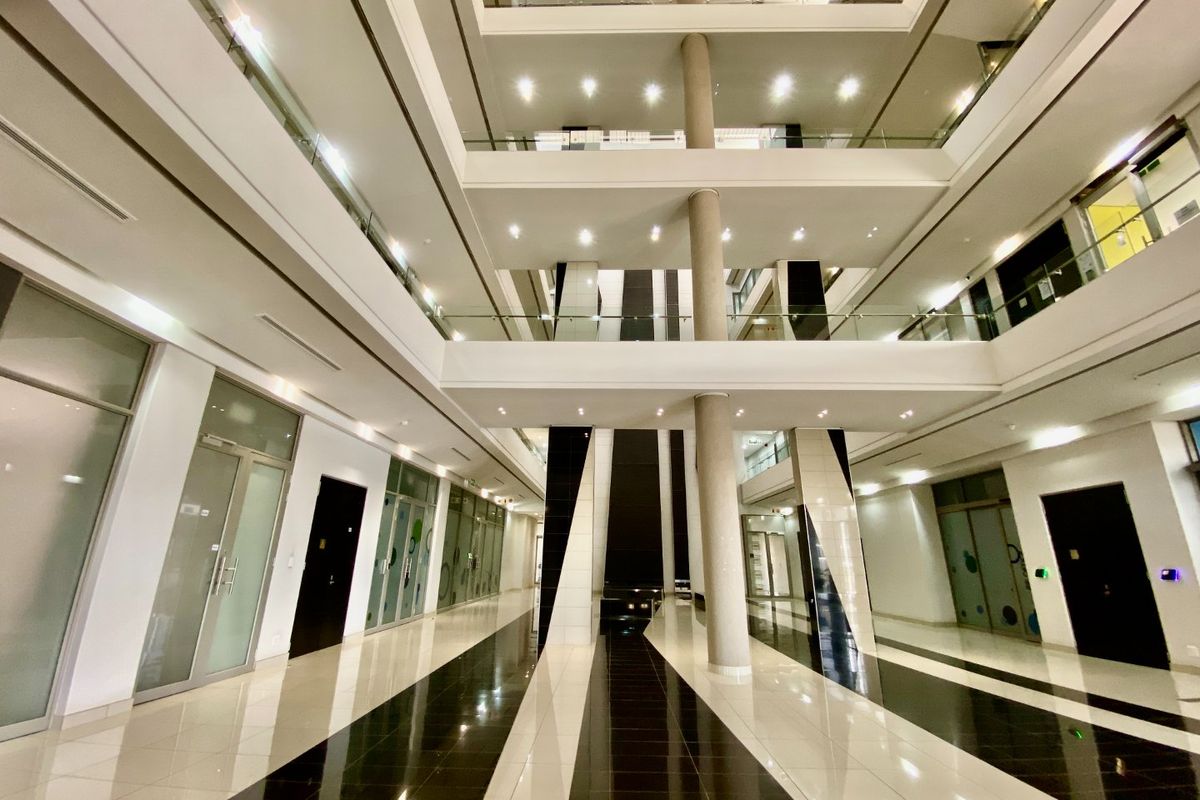 Spacious modern commercial atrium featuring high ceilings, glass balustrades, polished tiled flooring, and multiple glass-partitioned office entrances.