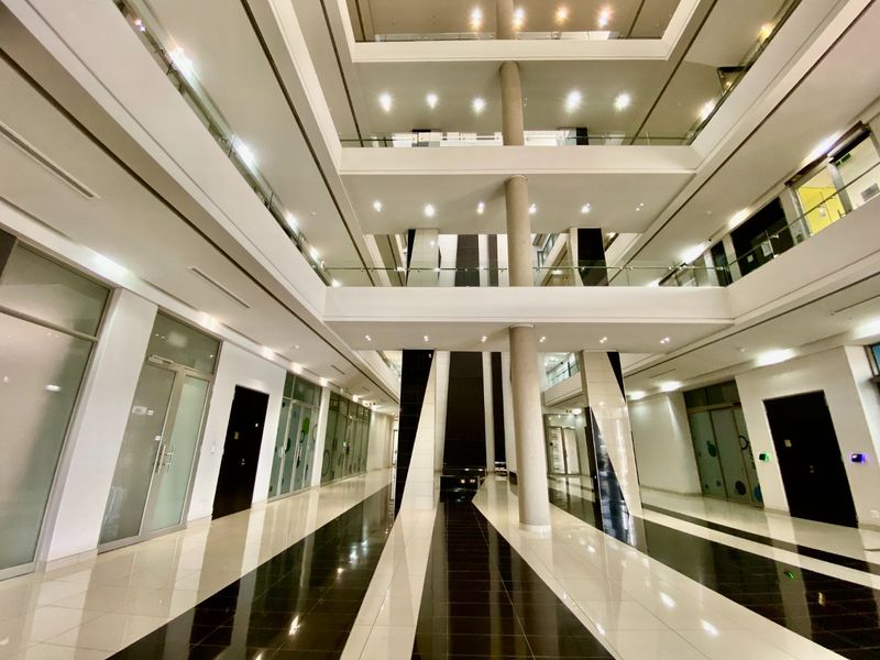 Spacious modern commercial atrium featuring high ceilings, glass balustrades, polished tiled flooring, and multiple glass-partitioned office entrances.