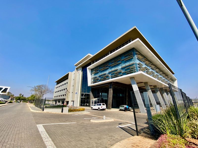 Modern commercial office building featuring a striking glass facade, cantilevered concrete design, and integrated sun louvers, set within a paved professional business park environment.