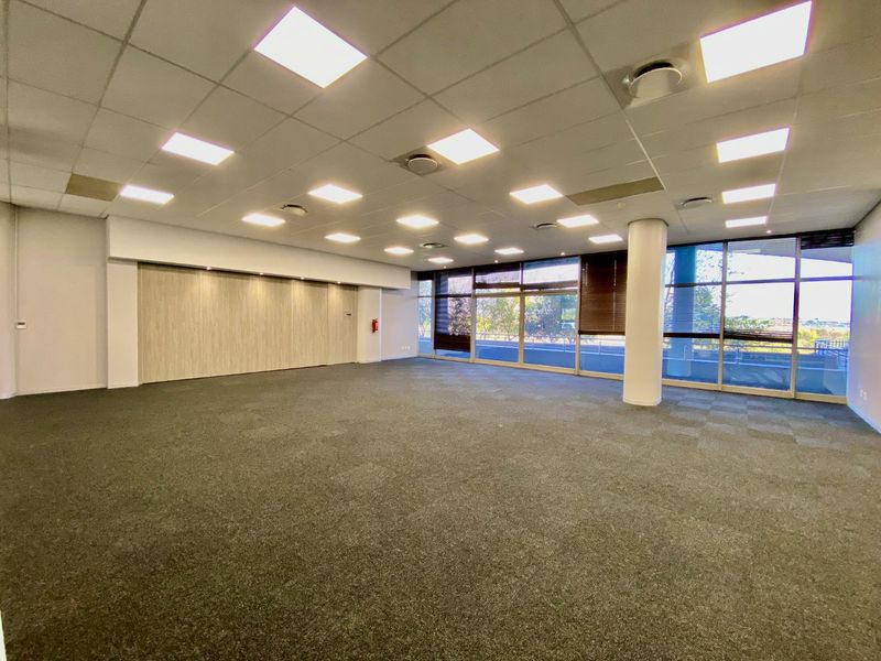 Spacious open-plan commercial office area featuring large windows, carpeted flooring, suspended ceilings with recessed lighting, and a flexible sliding partition wall.