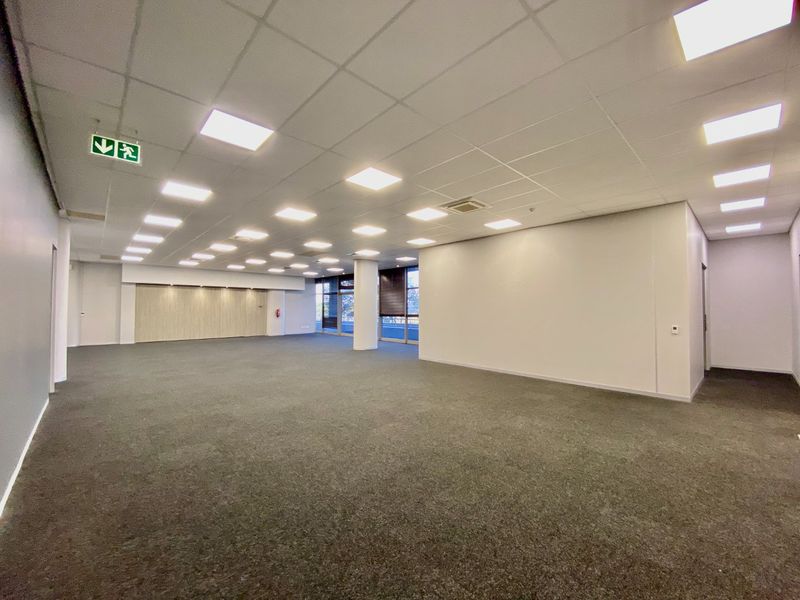A spacious, open-plan commercial office area featuring a suspended ceiling with recessed lighting, carpeted flooring, and a central structural column.