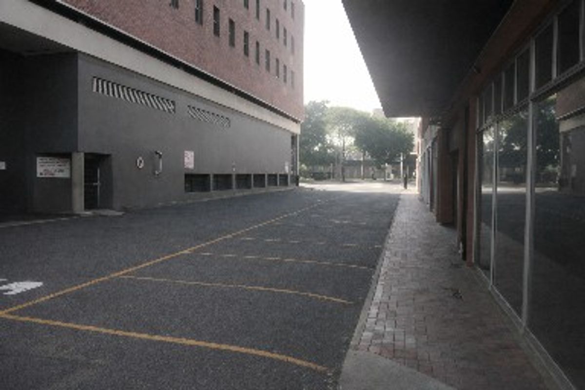Commercial exterior view showing an asphalt parking area, brick paved walkway, and large glass storefront windows under a covered overhang.