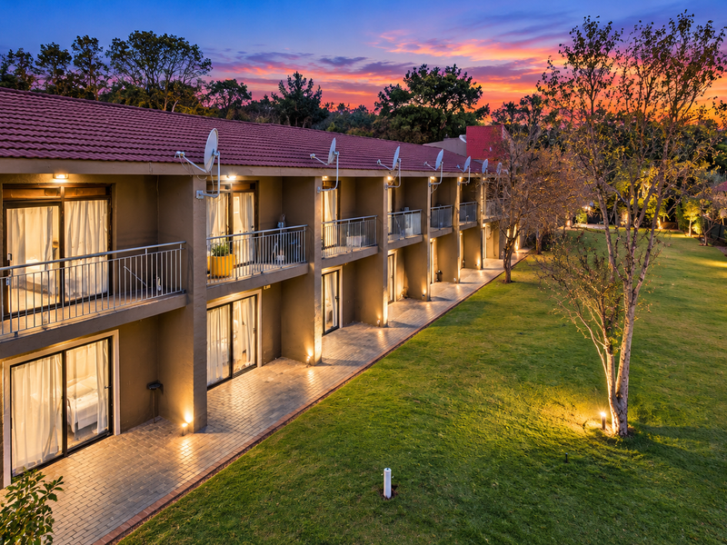 A well-maintained commercial property exterior featuring a long balcony with metal railings, sliding glass doors, and a spacious landscaped garden at dusk.