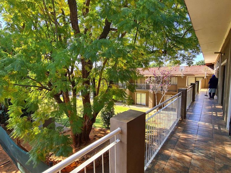 A spacious covered balcony featuring tiled flooring and a metal balustrade, overlooking the landscaped garden and surrounding property grounds.