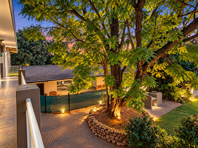 A well-maintained rear exterior area featuring a tiled walkway, stone-walled garden bed, mature tree, and paved patio space under a sunset sky.