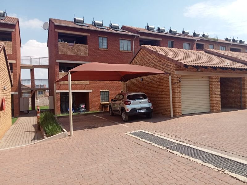 Exterior view of a face brick apartment complex featuring a paved driveway, covered carport parking, and roof-mounted solar water heating systems.
