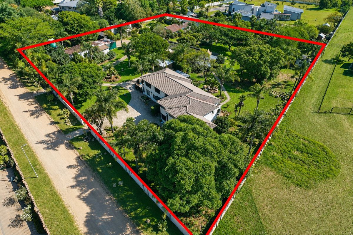 Aerial view of a residential property featuring a large landscaped garden, mature trees, paved driveway, and a tiled roof dwelling within a fenced perimeter.