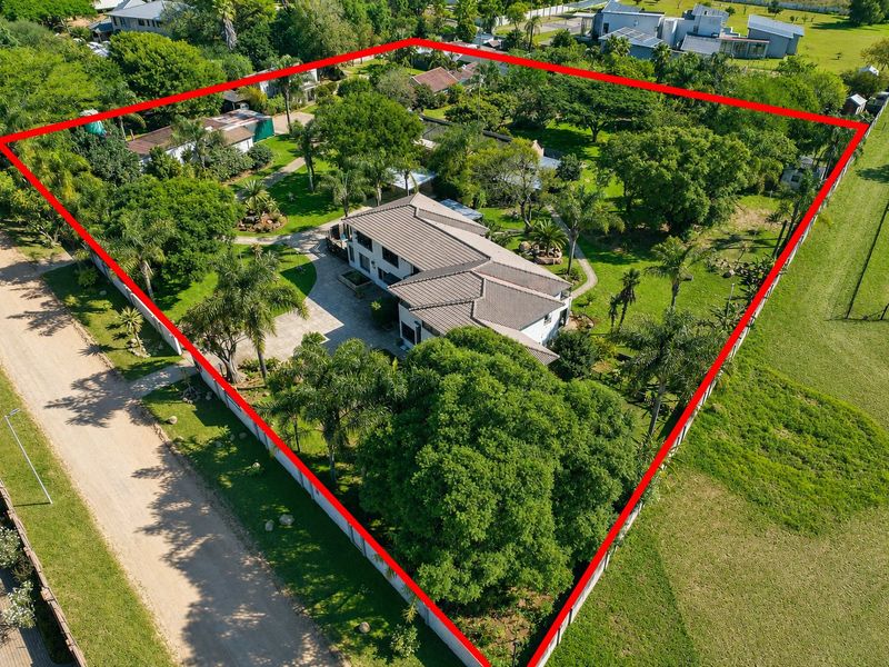 Aerial view of a residential property featuring a large landscaped garden, mature trees, paved driveway, and a tiled roof dwelling within a fenced perimeter.