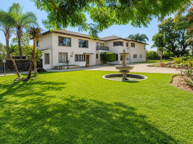 Spacious rear exterior view of the property featuring a well-maintained lawn, stone fountain, paved driveway, and established garden landscaping.