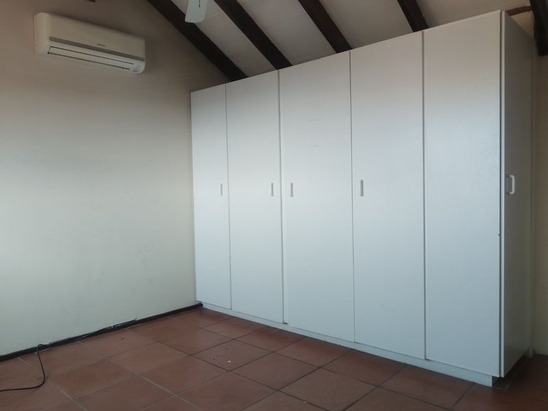 An empty bedroom featuring built-in white cupboards, exposed timber ceiling beams, tiled flooring, and a wall-mounted split-system air conditioning unit.