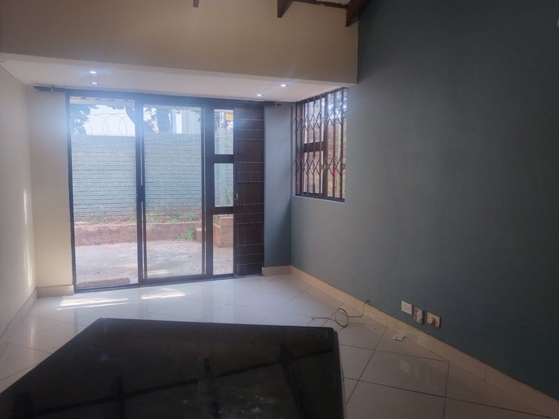 An empty living room featuring tiled floors, recessed lighting, exposed timber beams, and a sliding glass door with security gate leading to the exterior.