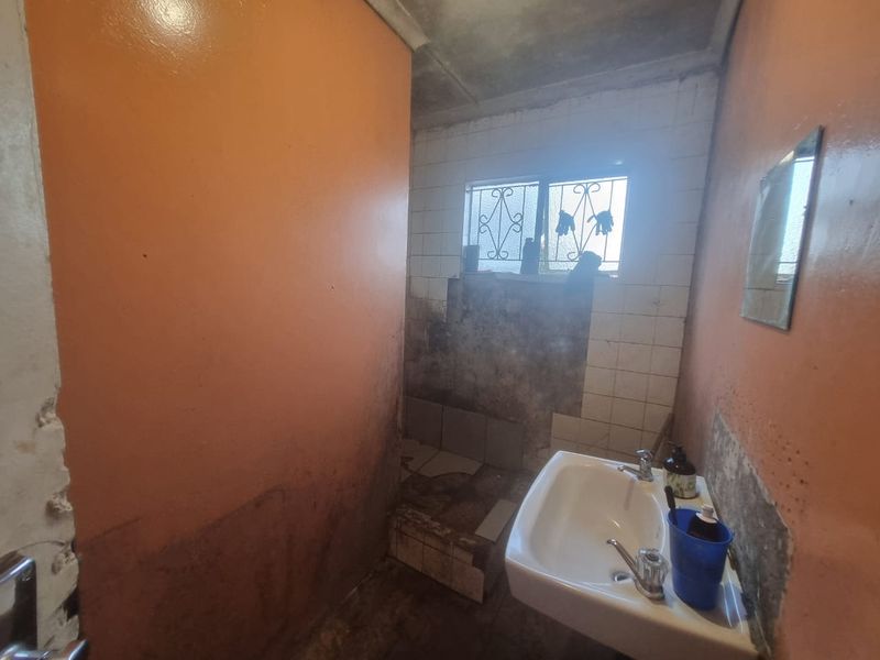 A small bathroom featuring a wall-mounted basin and window with security bars, currently requiring significant renovation and repair to wall finishes.