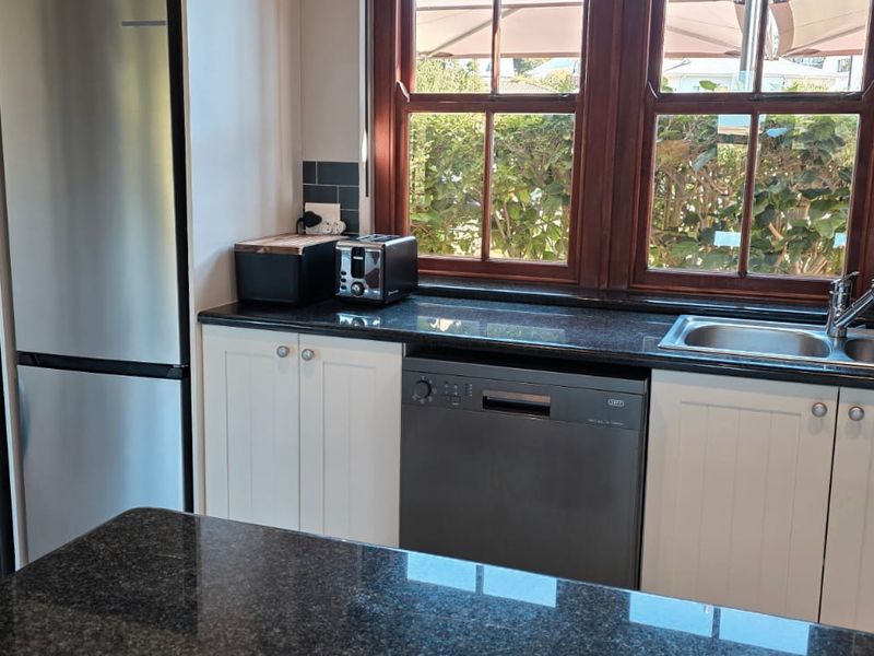A functional kitchen featuring granite countertops, white cabinetry, a stainless steel sink, and integrated dishwasher, with natural light from timber-framed windows.