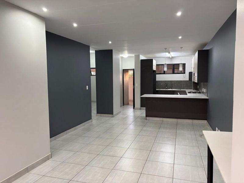 Modern open-plan living and kitchen area featuring tiled floors, dark cabinetry, a breakfast bar, and recessed lighting in a clean, vacant space.