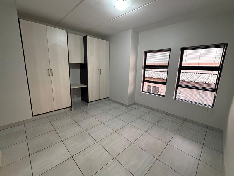 A clean, empty bedroom featuring neutral tiled flooring, built-in cupboards with an integrated desk space, and dual windows with fitted venetian blinds.