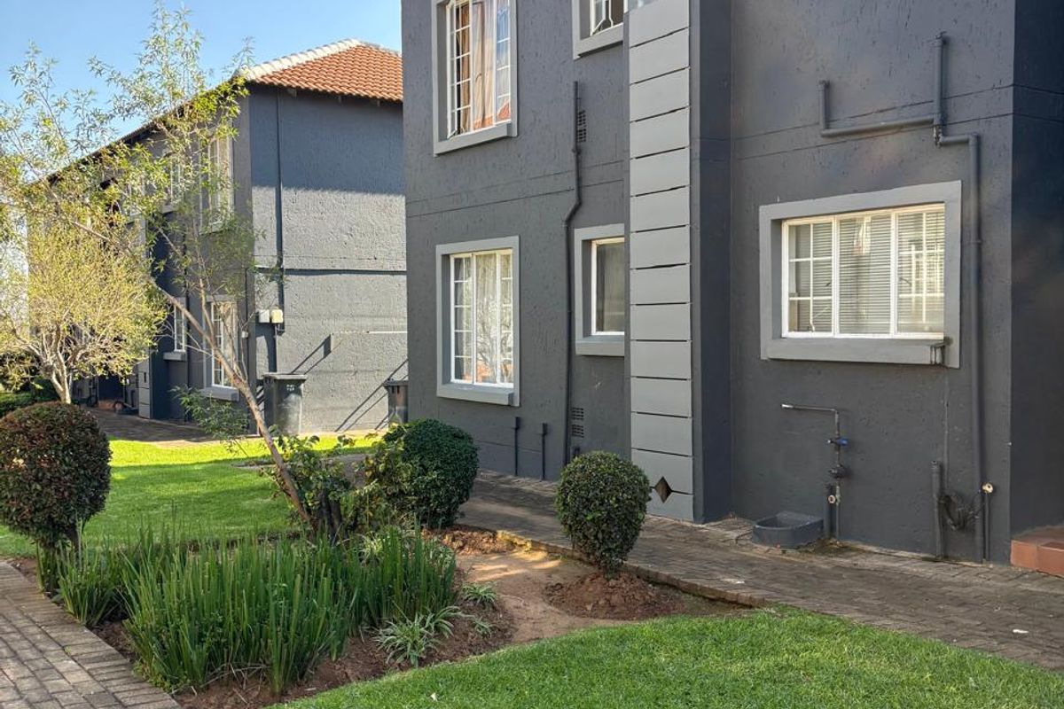 A view of the rear exterior of the apartment building, featuring a well-maintained garden, paved walkways, and dark-painted facade with casement windows.