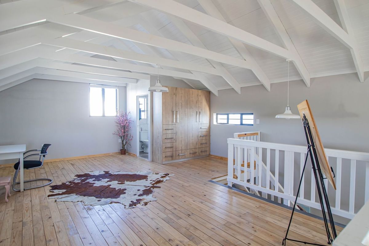Spacious loft-style bedroom featuring exposed white timber beams, natural wood flooring, and integrated built-in cabinetry with an adjoining en-suite bathroom.