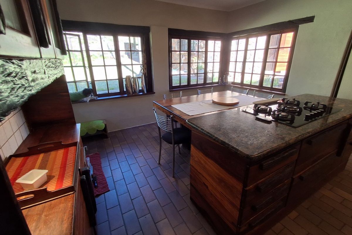 A rustic kitchen featuring wooden cabinetry, a central island with a gas hob, stone countertops, and tiled flooring with natural light from timber-framed windows.