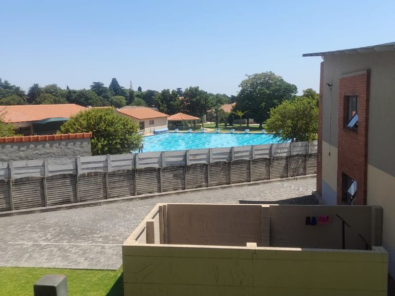 An elevated view overlooking a communal swimming pool area, paved driveway, and landscaped lawn within the apartment complex grounds.