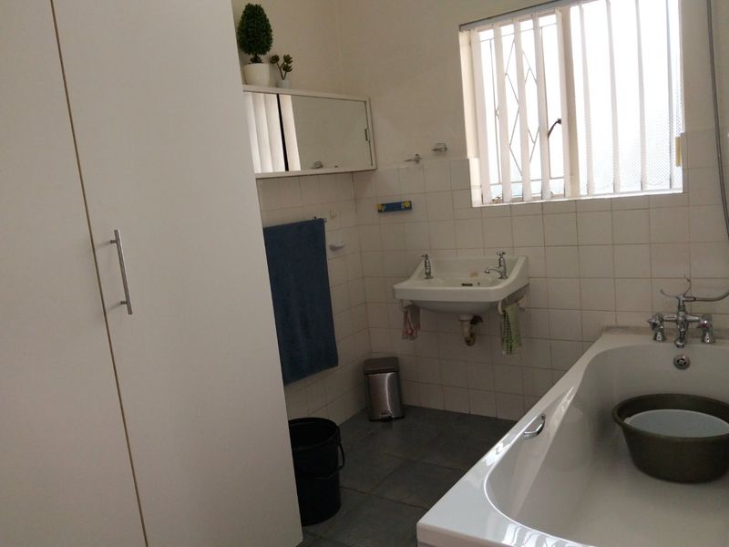 A functional bathroom featuring a bathtub, wall-mounted basin, built-in storage cupboard, and tiled finishes with security bars on the window.