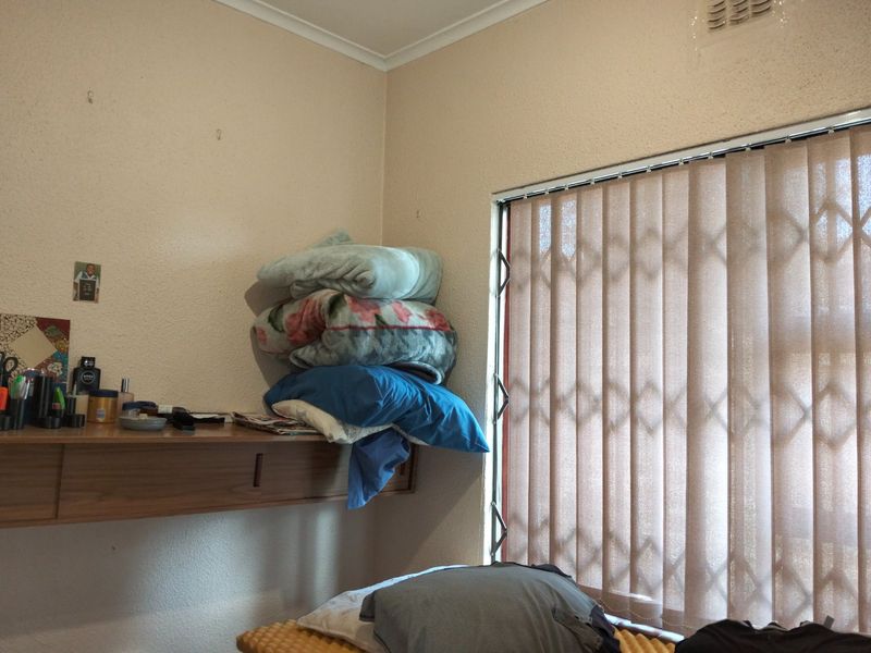 A bedroom corner featuring a built-in floating shelf, vertical window blinds, and a security gate, showing signs of wear on the wall surfaces.