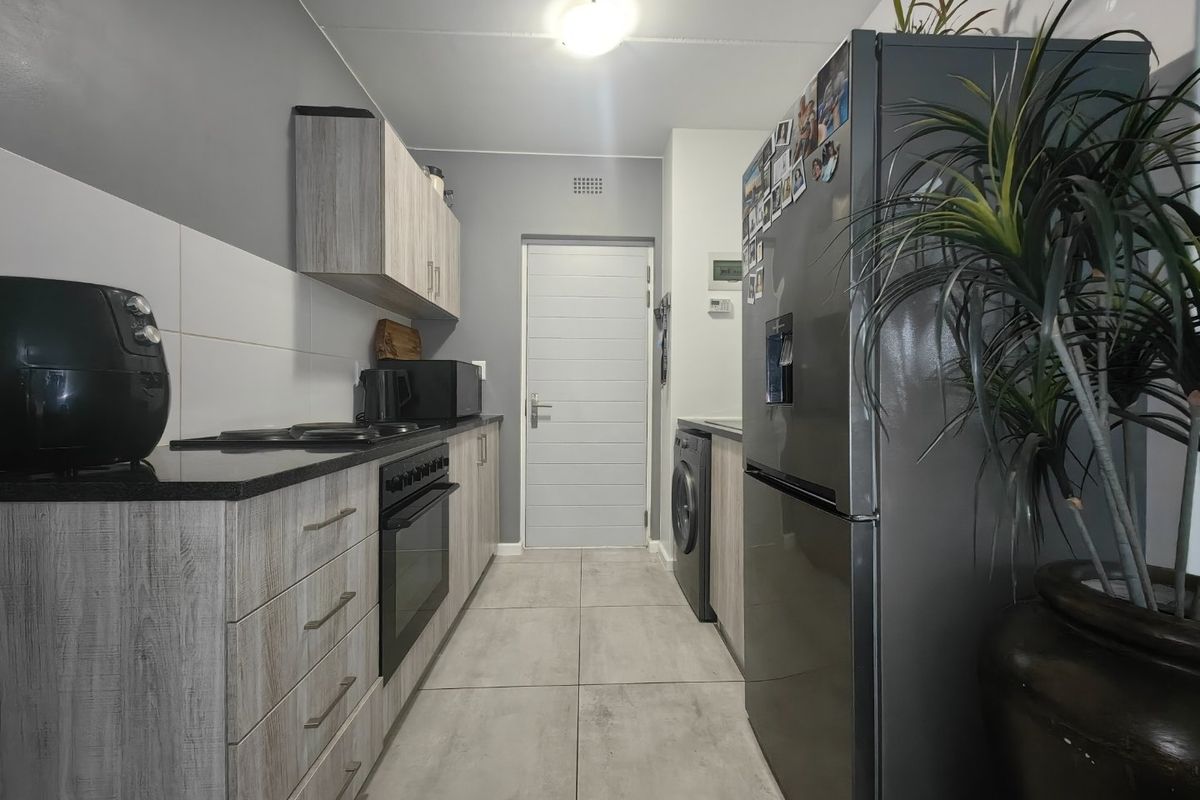 Modern galley-style kitchen featuring light wood-grain cabinetry, dark stone countertops, integrated oven and hob, and neutral floor tiling.