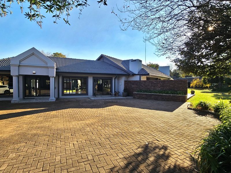 A well-maintained front exterior featuring a spacious paved driveway, a covered entrance portico with columns, and a landscaped garden with a brick feature wall.