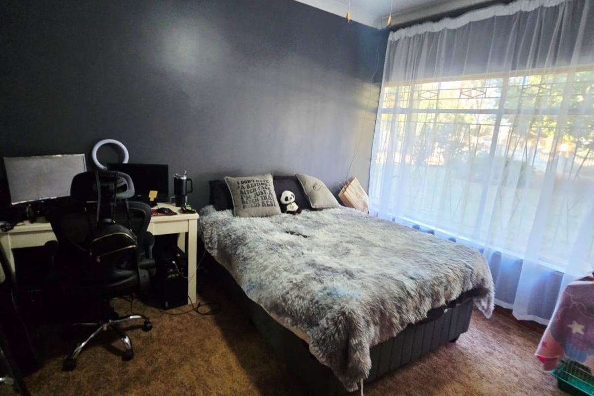 A bedroom featuring a ceiling fan, large window with natural light, and dark accent wall, currently utilized as a combined sleeping and workspace area.
