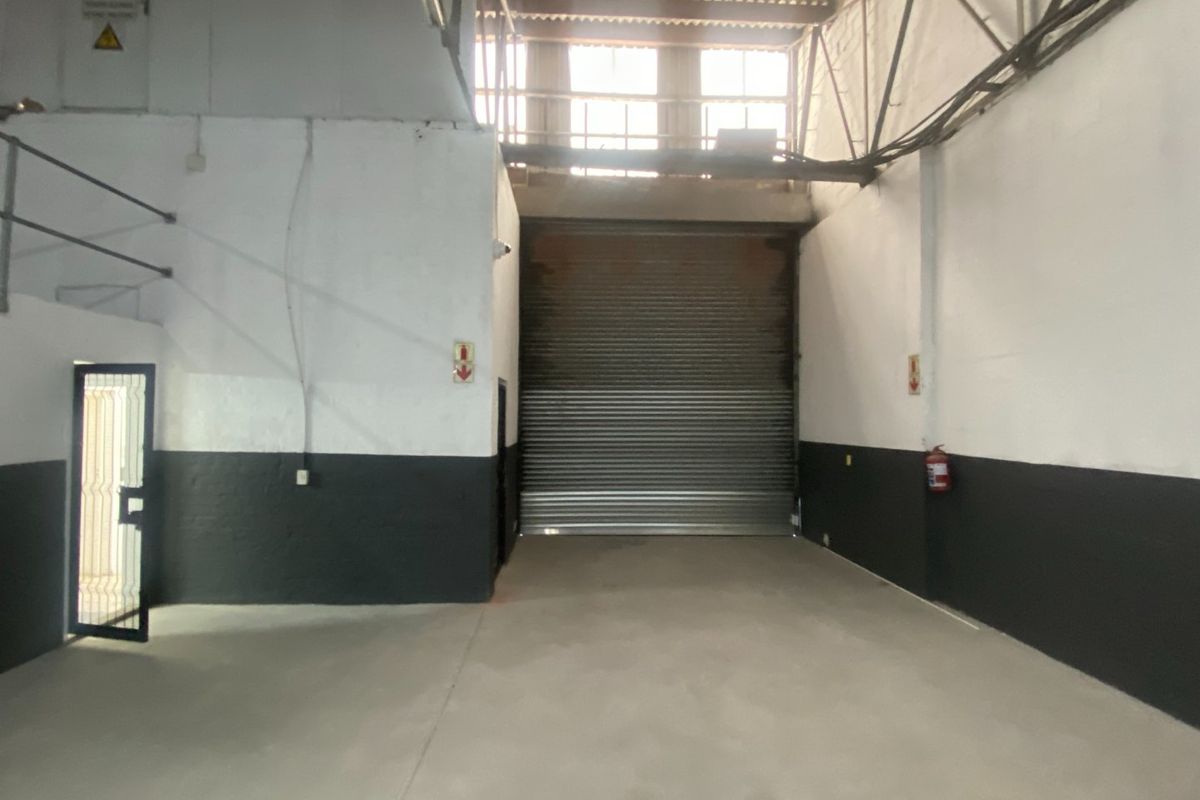 Industrial warehouse space featuring high ceilings with exposed steel trusses, concrete flooring, and a large roller shutter door for vehicle or goods access.