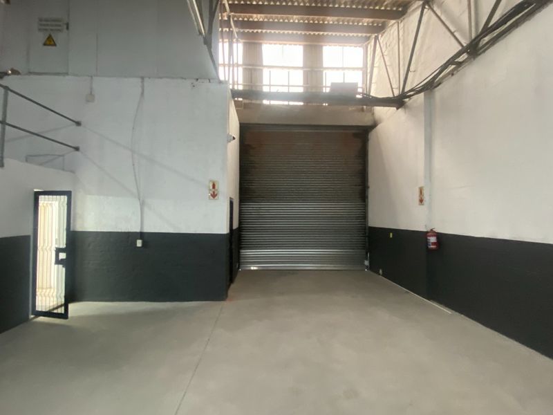 Industrial warehouse space featuring high ceilings with exposed steel trusses, concrete flooring, and a large roller shutter door for vehicle or goods access.