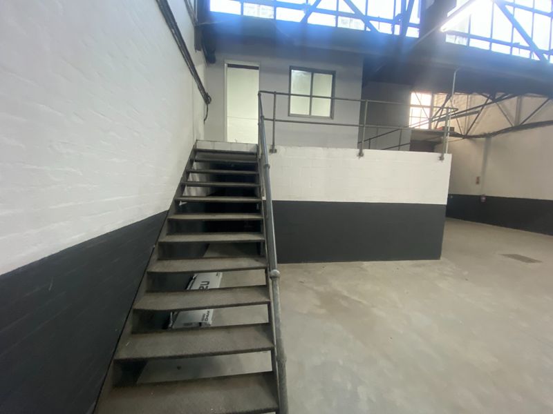 Industrial utility space featuring a metal staircase, exposed steel roof trusses, and painted brick walls, offering a functional open-plan layout.